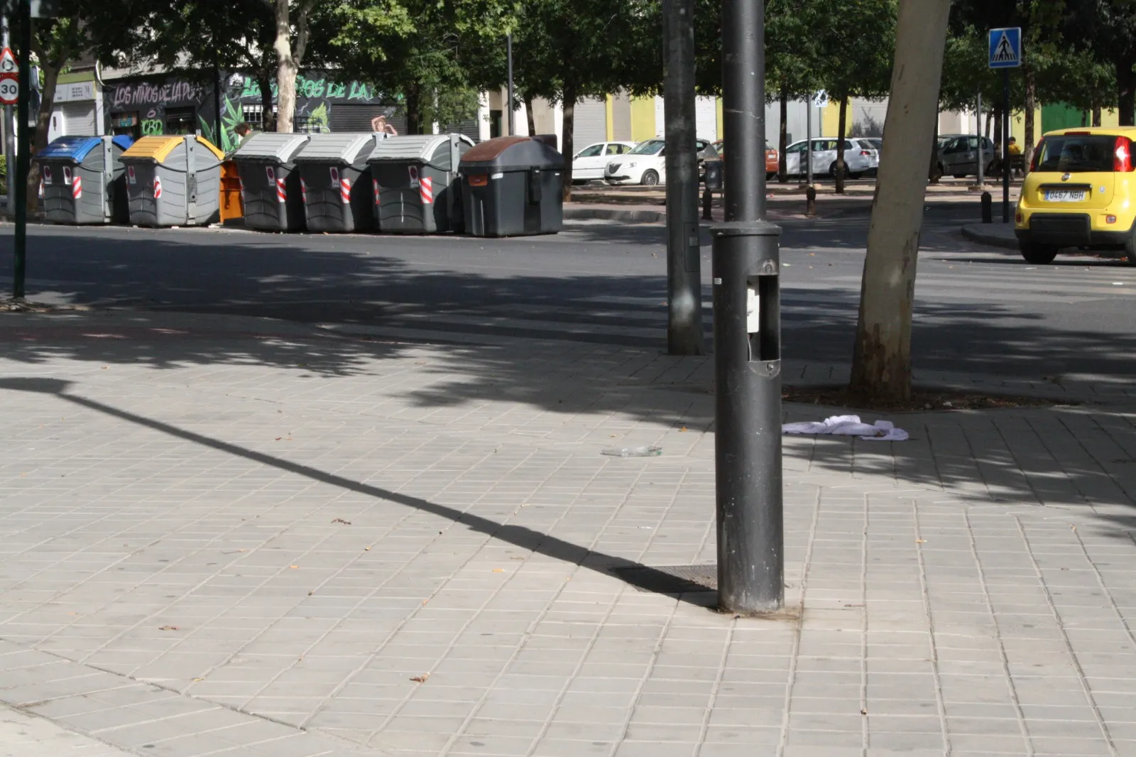 Acera junto a una calle con contenedores; bajo un árbol hay ropa de alguien en el suelo